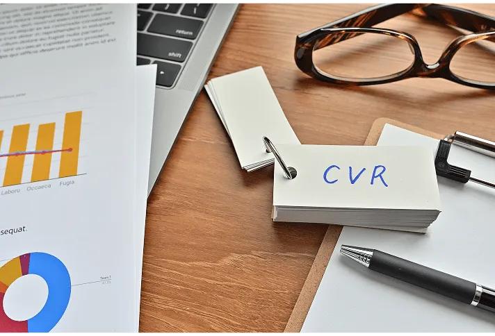 木製デスクに置かれたクリップボードに「CVR」と書かれたメモ、周りにメガネ、ペン、ノートパソコンが配置されている