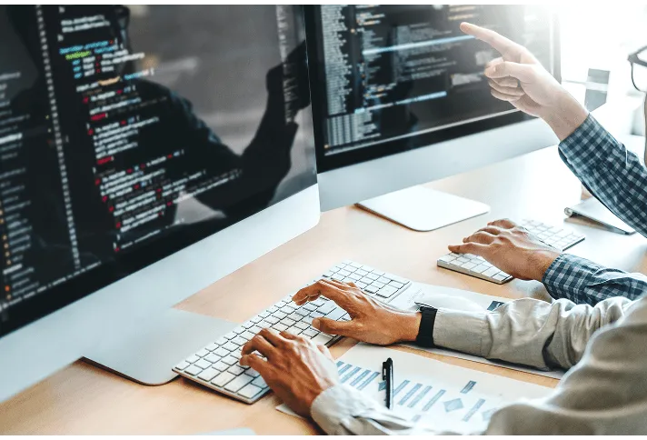 デュアルモニターにプログラムコードを表示させ、白いキーボードでコーディング作業をしている開発者の手元