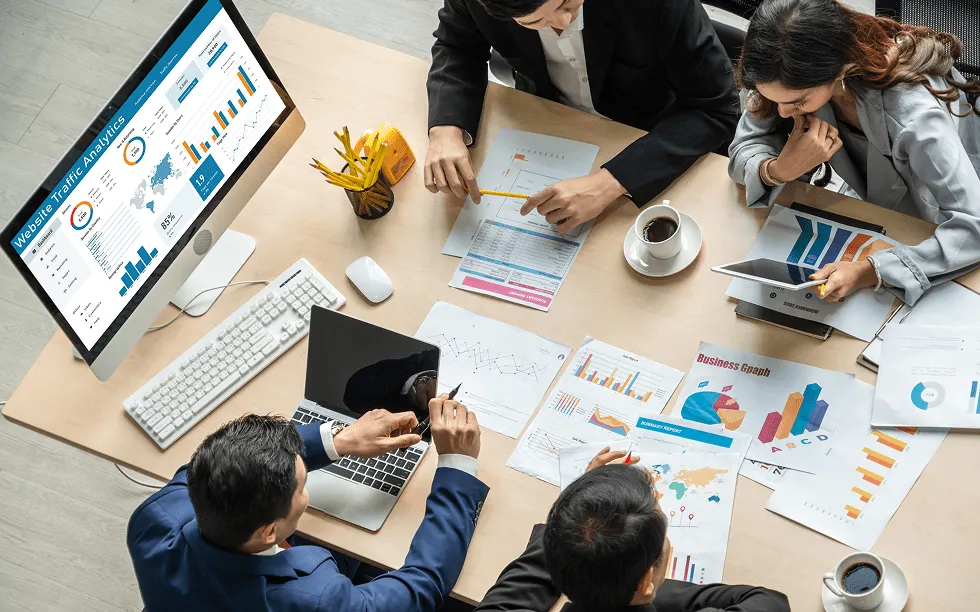 デスクトップモニターにWebサイトの分析データを表示し、4人のビジネスチームが様々なグラフ資料を囲んで分析作業をしている俯瞰図
